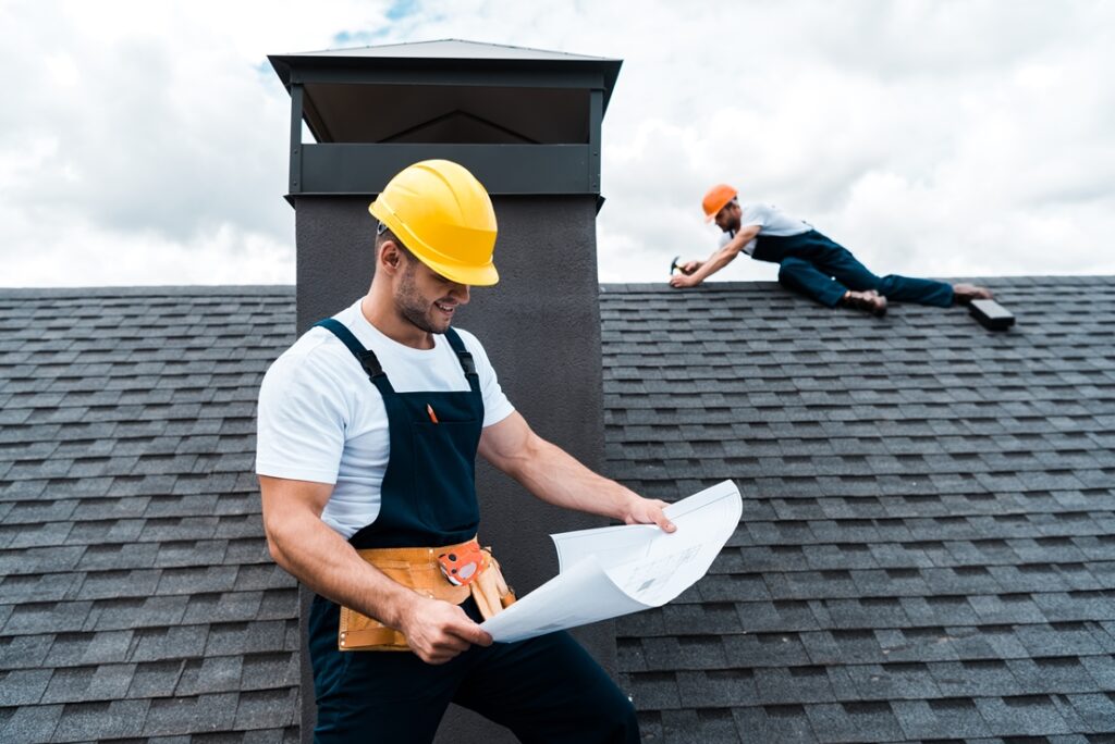 Selective focus of handsome builder in helmet holding blueprint near colleague repairing roof