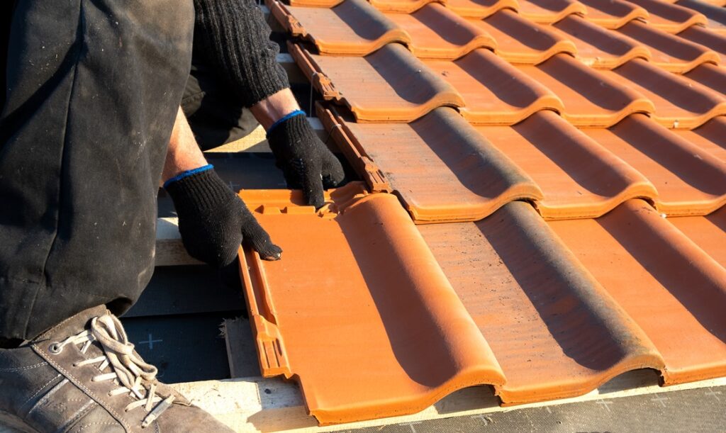Closeup of worker hands installing yellow ceramic roofing tiles mounted on wooden boards covering residential building roof under construction.